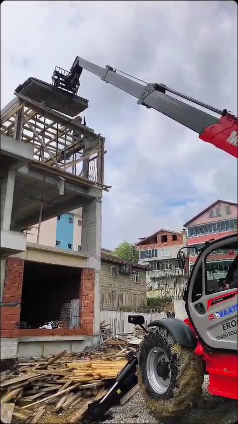 Yamaç yerleşimde çatı makasına kereste taşıyan teleskopik yükleyici, önde hurda kereste yığını
